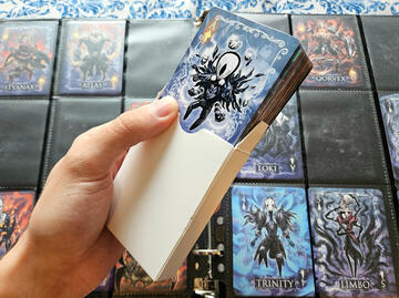 Photo of a full deck of Hollowframe playing cards sticking out of its blank white tuck box, in front of an opened binder with remaining cards from TennoCon 2025.